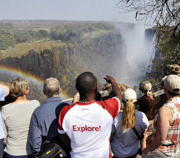 People gather at a scenic overlook, pointing towards a large waterfall. A rainbow spans the misty gorge. One person wears a shirt with "Explore!" printed on it. Dense greenery surrounds.